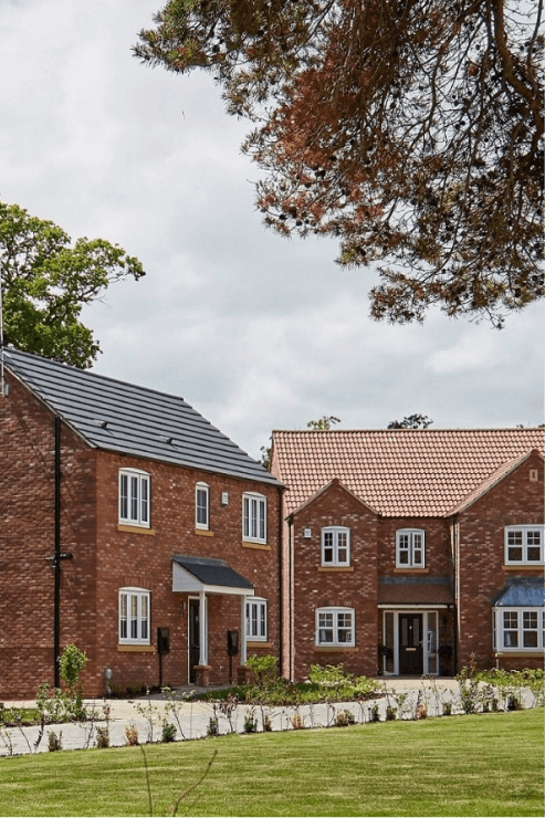 Two detached red brick new-build houses with tiled roofs and white window frames set in a green landscaped development