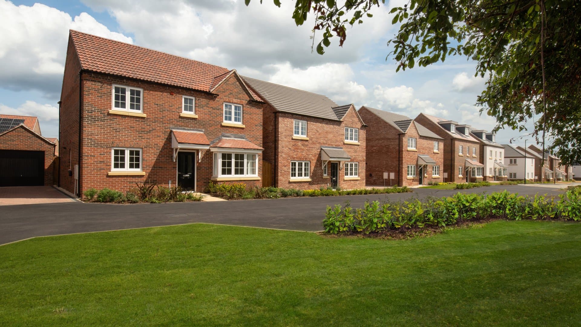 A row of modern detached brick houses with a paved road, manicured lawns and trees under a blue sky with clouds