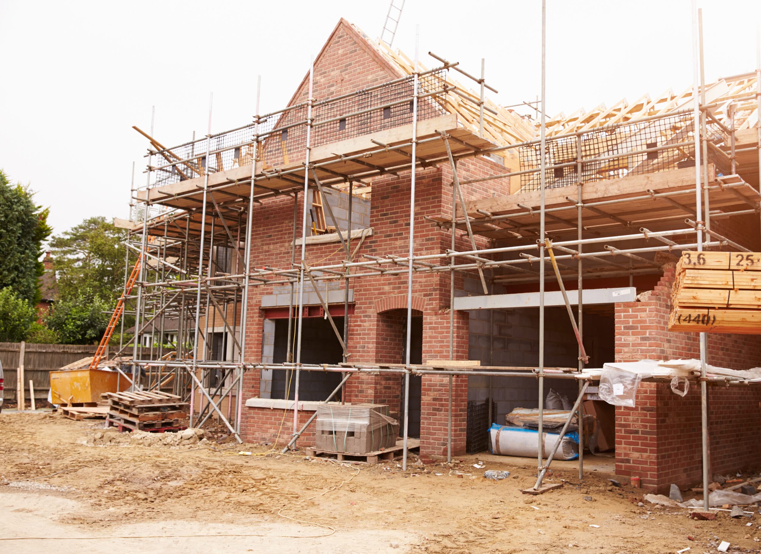 Brick house under construction with scaffolding, wooden roof trusses, and building materials on site