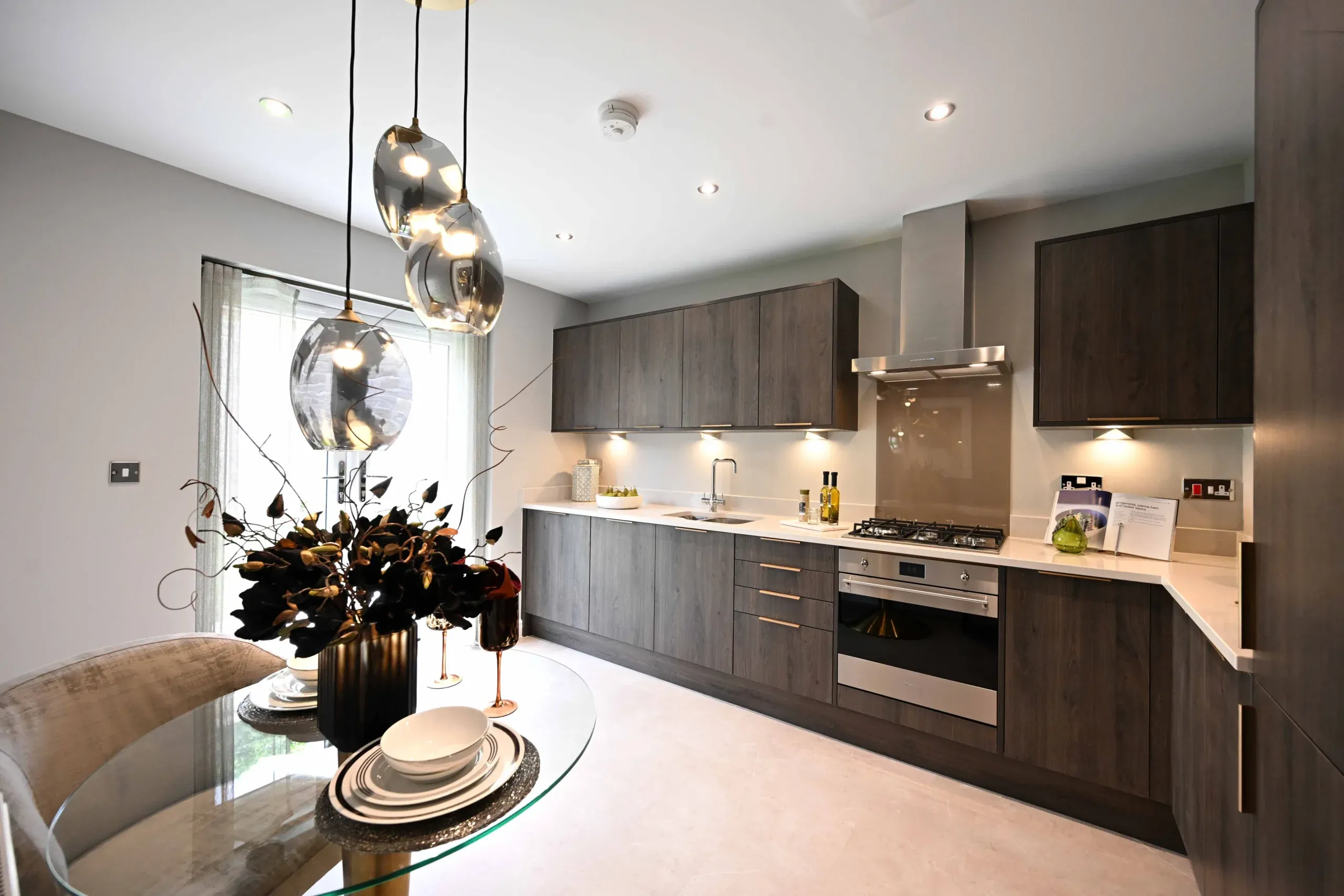 Modern kitchen with dark wood cabinetry, integrated appliances, and a glass dining table with pendant lighting