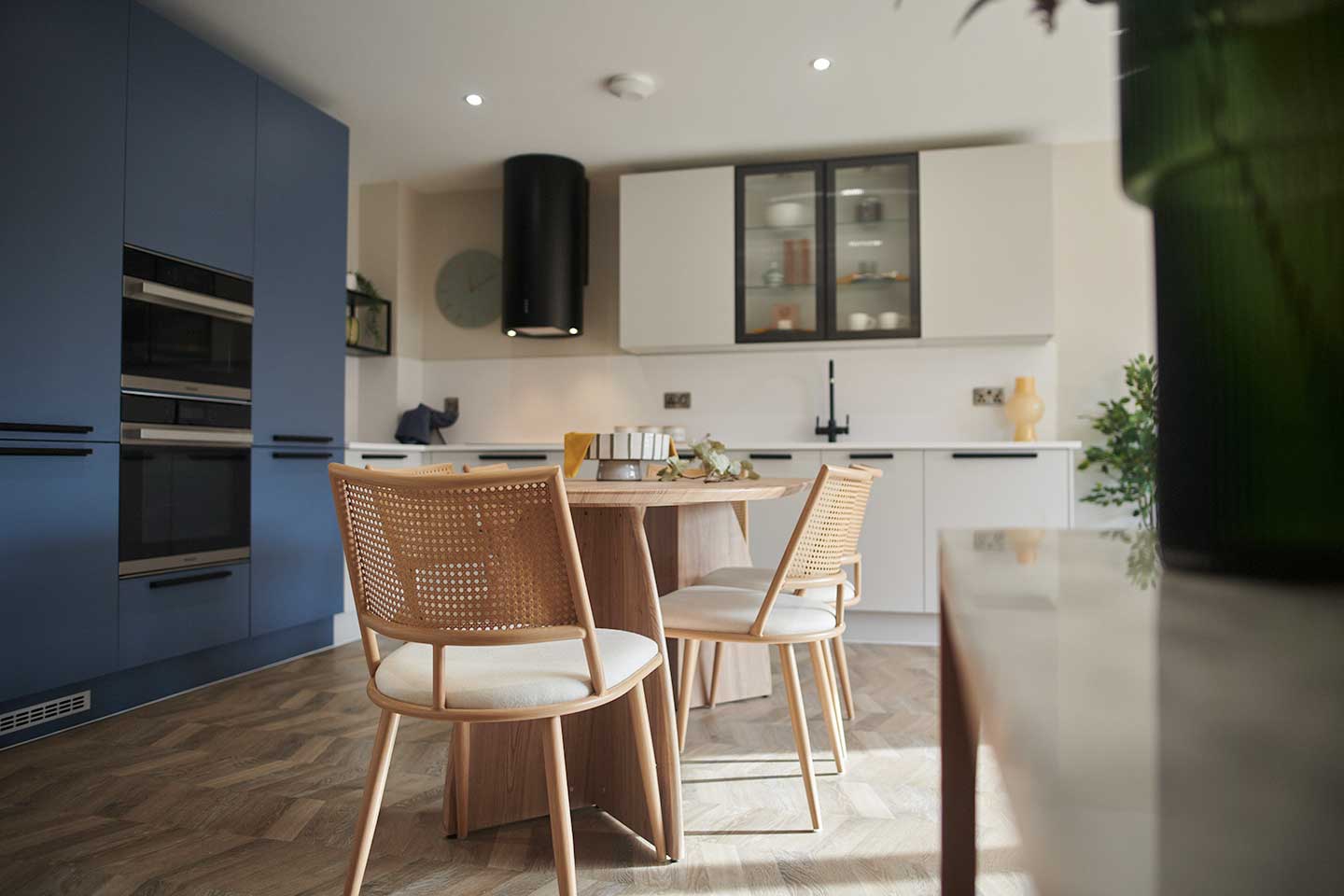 Modern kitchen with blue units, white cupboards, and a central wooden dining table with cane-back chairs on parquet floo