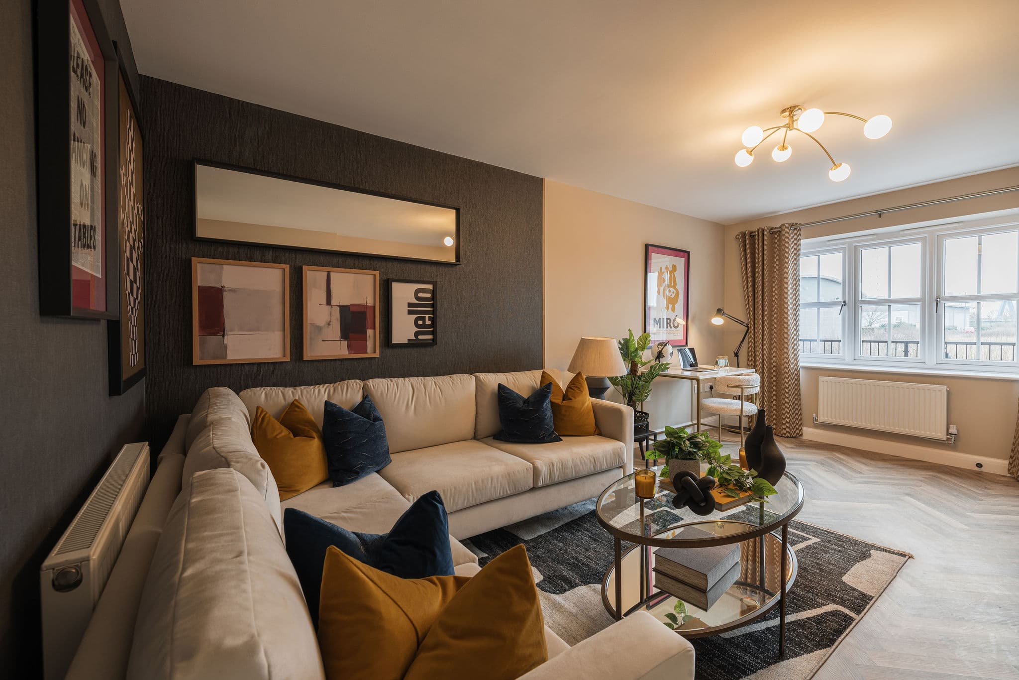 Modern living room with a beige corner sofa, dark grey feature wall, glass coffee table, and a small study area by windo