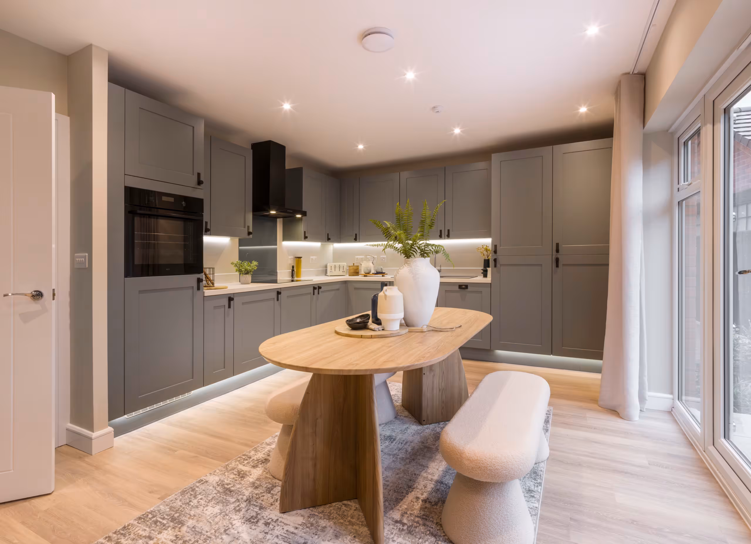 Modern kitchen with sage green cabinets, light wood dining table with bench seating, and glass doors leading outside