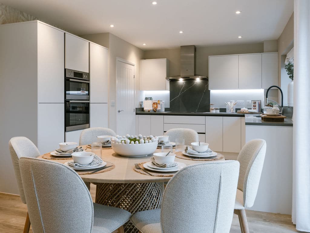 Modern open-plan kitchen and dining area with a round wooden table, white upholstered chairs, and integrated appliances