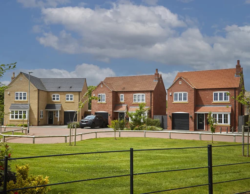 Row of three new-build detached brick houses with garages, set behind a green common area and metal fencing