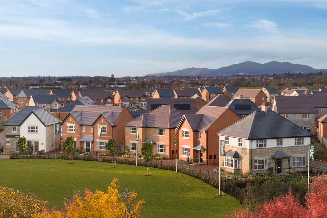 Drone view of a modern UK housing development with detached brick homes, green lawns, and distant hills.